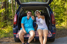 The traveler is sitting in the trunk of a car laughing and pointing a finger at the camera. A man and a woman travel in their minivan. People are resting from a long trip on a forest road.