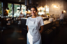 A Beautiful, Smiling Young Brunette Chef In A White T-shirt And Blank White Apron Stands In The Middle Of The Restaurant With A Cozy Interior With Lamps On. Mock-up For Design. Blank Template.