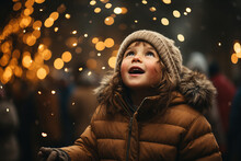Child In Winter Holiday Event On Festive Lights Background. Kid Wear Sweater And Hat Looking Up And Stands In Wonder Before A Lit Huge Christmas Tree Under The Night Sky. Magic Moment.