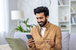 © Liubomir - Young joyful man sitting on sofa at home, using application on phone for online shopping in online store, Indian man smiling contentedly holding bank credit card.