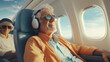 A happy senior man with white hair and sunglasses listens to music with headphones while traveling by plane. He enjoys his retirement and the freedom of being a pensioner. Happiness in old age.