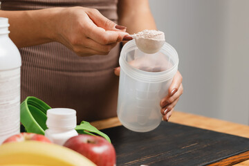 Wall Mural - Athletic woman in sportswear with measuring spoon in her hand puts portion of whey protein powder into a shaker on wooden table with amino acid white capsules, bananas and apple, making protein drink