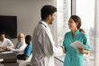 © fizkes - Positive young medical colleagues shaking hands in meeting room with team talking in background. Practitioner doctor man giving handshake to happy surgeon woman on conference