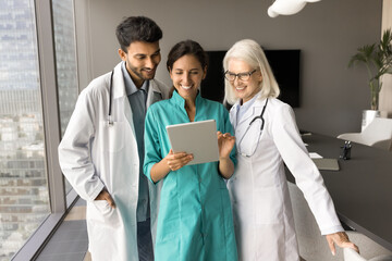 Wall Mural - Happy multiethnic clinic medical staff in uniforms using tablet computer together, talking, laughing, discussing healthcare online application, enjoying online communication, modern technology