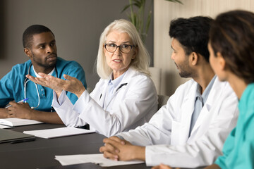 Poster - Serious senior head doctor talking to diverse hospital staff at meeting table, speaking, giving instructions, explaining medical problem. Medical expert, mentor training younger interns