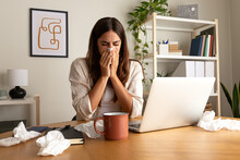 Sick woman working from home office. Caucasian female blowing nose with tissue while working with laptop.