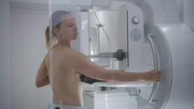 Caucasian adult woman stands topless in hospital radiology room. Female patient undergoing mammography screening checkup using digital mammograph. Breast cancer prevention. Modern clinic equipment.