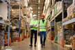 © alfa27 - two men engineers in safety helmets and green uniforms walking among shelves with goods in warehouse talking, checking goods. logistic and business export, distribution and inventory concept