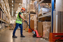 Male Staff Worker In Warehouse Uses Hand Pallet Stacker To Transport Goods, Alone, Dressed In Working Clothes And Safety Hard Hat.Skilled Warehouse Employee Pushing Manual Pallet Jack