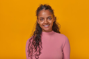 Young optimistic cute Indian woman teenager with black curly hair looking at camera and smiling posing for student album dressed in casual style stands in orange studio. Portrait ethnic girl