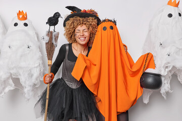 Wall Mural - Positive optimistic woman embraces orange ghost holds broom with crow on it smiles broadly keeps eyes closed isolated over white background surrounded by ghosts. Halloween celebration concept