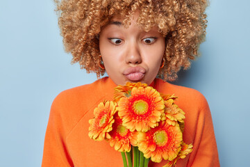 Wall Mural - People emotions concept. Indoor waist up of young surprised African american woman wearing orange sweater holding and looking at bouquet of gerbera flowers standing in centre on blue background