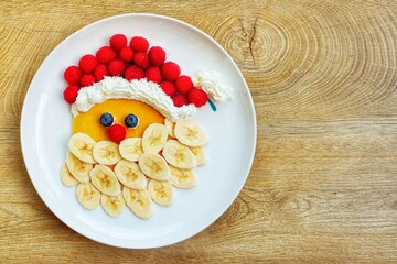 Wall Mural - Santa claus pancake made it from pancake,bananas,blueberries,raspberries and whipped cream on plate with wooden background.Creative art food idea for kids Christmas breakfast.Top view.Copy space

