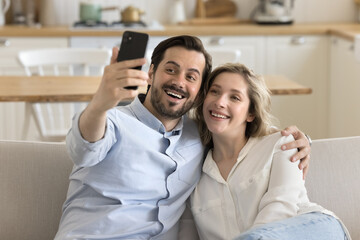 Wall Mural - Beautiful millennial couple resting on sofa staring at cell phone screen, make selfie picture, calling to friends using videocall application spend weekend together at home. Leisure using modern tech