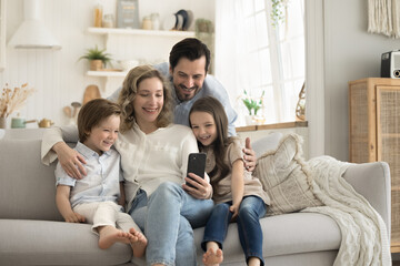 Wall Mural - Happy family spend time in studio apartment sit on couch, smiling mother hold mobile phone take selfie photo together for memory, kids have great time with parents, make online call with grandparents