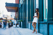 Beautiful American girl stands tall near a skyscraper in the city. Black woman wears stylish white dress and heels outdoors 