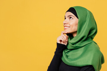 Wall Mural - Side view young fun arabian asian muslim woman wear green hijab abaya black clothes prop up on chin look aside isolated on plain yellow background. People uae middle eastern islam religious concept.