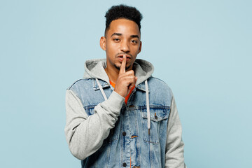 Wall Mural - Young fun secret man of African American ethnicity wear denim jacket orange t-shirt say hush be quiet with finger on lips shhh gesture isolated on plain pastel light blue background studio portrait.