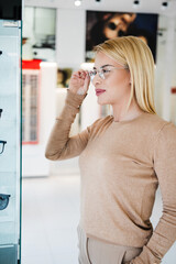 Poster - Beautiful and fashionable woman choosing eyeglasses frame in modern optical store.