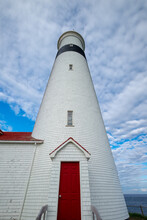 A Tall Circular Red And White Lighthouse With An Attached Light Keeper's House. The Sky Is Very Dramatic With Lots Of White Clouds And A Blue Sky Underneath.  There's Grass In Front Of The Lighthouse.
