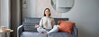 Relaxation and patience. Smiling young asian woman in cozy room, sitting on sofa and meditating, doing yoga mindfulness training