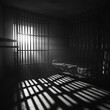 Somber empty room cell with shadows of bars reflected on the floor. Soft light entering from a small window inside the prison cell. Black and white Jail 