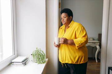 Sad upset lonely plus size elderly retired female of african american ethnicity standing in front of window with white cup of hot drink in hands, looking at city streets, missing her grandchildren