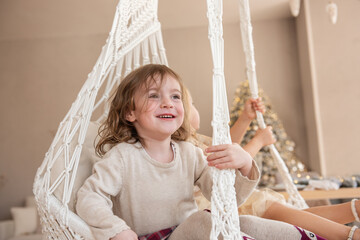 Two little girls ride on a macrame swing at home near the Christmas tree. Sisters have fun, fooling around on vacation. Funny girlfriends having a happy time in a Scandinavian trendy interior