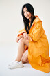 Pretty asian girl in bright yellow raincoat posing on white studio background