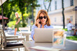 © sepy - Cheerful smiling businesswoman using laptop and cellphone and working at outdoor cafe terrace