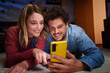 © CarlosBarquero - Happy nice Caucasian couple using cell phone smiling lying on carpet in living room at home at night. Young adult partner relationship enjoying mobile together. Domestic life positive people indoor.