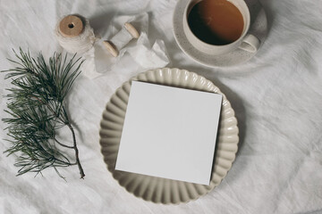 Wall Mural - Christmas stationery. Blank square greeting card, invitation mockup, ceramic plate. Silk ribbon and pine tree branches. White linen tablecloth. Cup of coffee, winter breakfast. Festive holiday flatlay