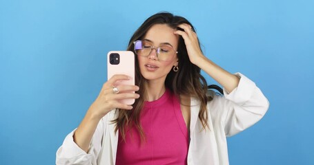 Wall Mural - The girl stares at the phone and fixes her hair. Female uses phone like mirror. Young girl make photo from hands with phone on blue background, look at camera and smiling, she wear eye glasses.