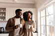 Happy dreamy African American young couple drinking hot beverages, holding cups, standing in kitchen