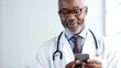 © GT77 - Elderly, smiling African American doctor in glasses and a white lab coat is looking at the screen of a phone that he's holding in his hands. Communication after work. Banner with copy space