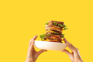 Female hands holding plate with tasty sandwich on yellow background, closeup