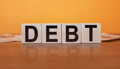 Wall Mural - DEBT - an inscription on wooden cubes on an orange background along with banknotes. Finance and wear concept.