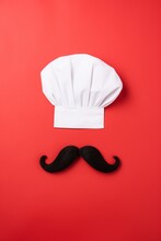 White Chef's Uniform Hat And Black Curled Ends Pointed Mustache. Minimal Concept Of Kitchen, Restaurant, Recipe. Symbol Or Icon Of Fine Dining Cuisine. Flat Lay Creative Retro Composition And Design