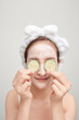 Beautiful young woman with facial mask on her face holding slices of fresh cucumber.