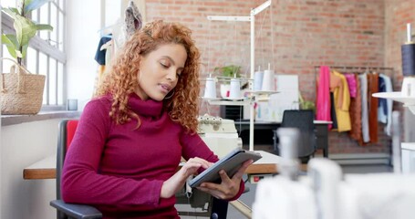 Wall Mural - happy biracial female fashion designer sitting and using tablet in studio, slow motion