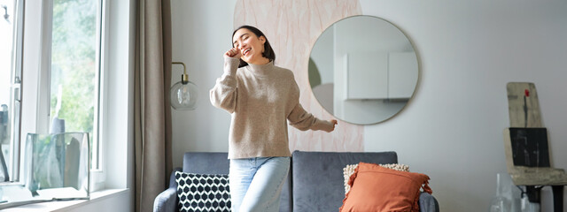 Wall Mural - Happy teen girl dancing at home, singing along music, using hand as microphone, enjoying happy day