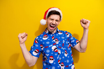 Poster - Photo of young funny guy yell raised fists up hooray funny print shirt santa claus sunglasses xmas eve isolated on yellow color background