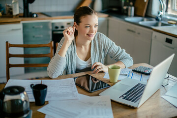 Wall Mural - Young woman using her laptop and going over bills and payments in the kitchen