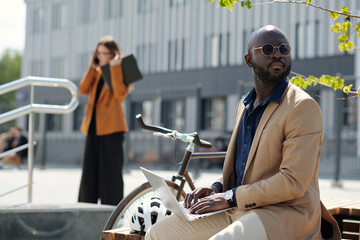 Wall Mural - Stylish young businessman in formalwear and sunglasses typing on laptop keyboard while sitting in front of camera against colleague