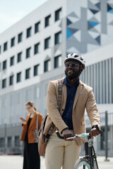 Poster - Happy young African American businessman in cycling helmet standing by his bicycle against modern architecture while riding to work