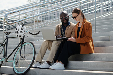Poster - Two young intercultural colleagues discussing data in document held by pretty Caucasian businesswoman while both sitting on staircase