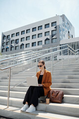 Wall Mural - Long shot of young modern businesswoman with laptop networking on staircase in urban environment against multi-storey office center