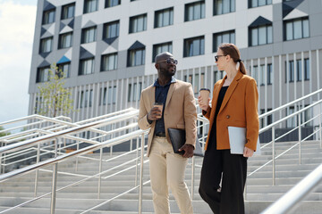 Wall Mural - Modern young employees in quiet luxury apparel talking to one another and having coffee while moving downstairs in urban environment