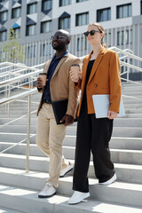Poster - Two young intercultural colleagues with cups of coffee moving downstairs in urban environment while one of them carrying folded laptop