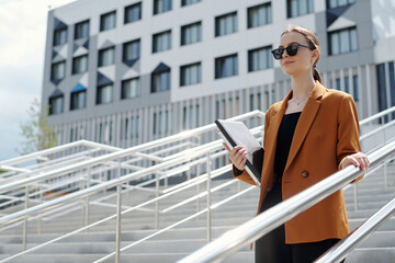 Wall Mural - Young confident female broker or entrepreneur carrying folders with financial papers while holding by railings and moving downstairs